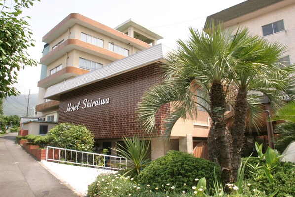 A hotel named "Hotel Shiraiwa" with a brick facade, a balcony, and surrounding greenery including palm trees and bushes.