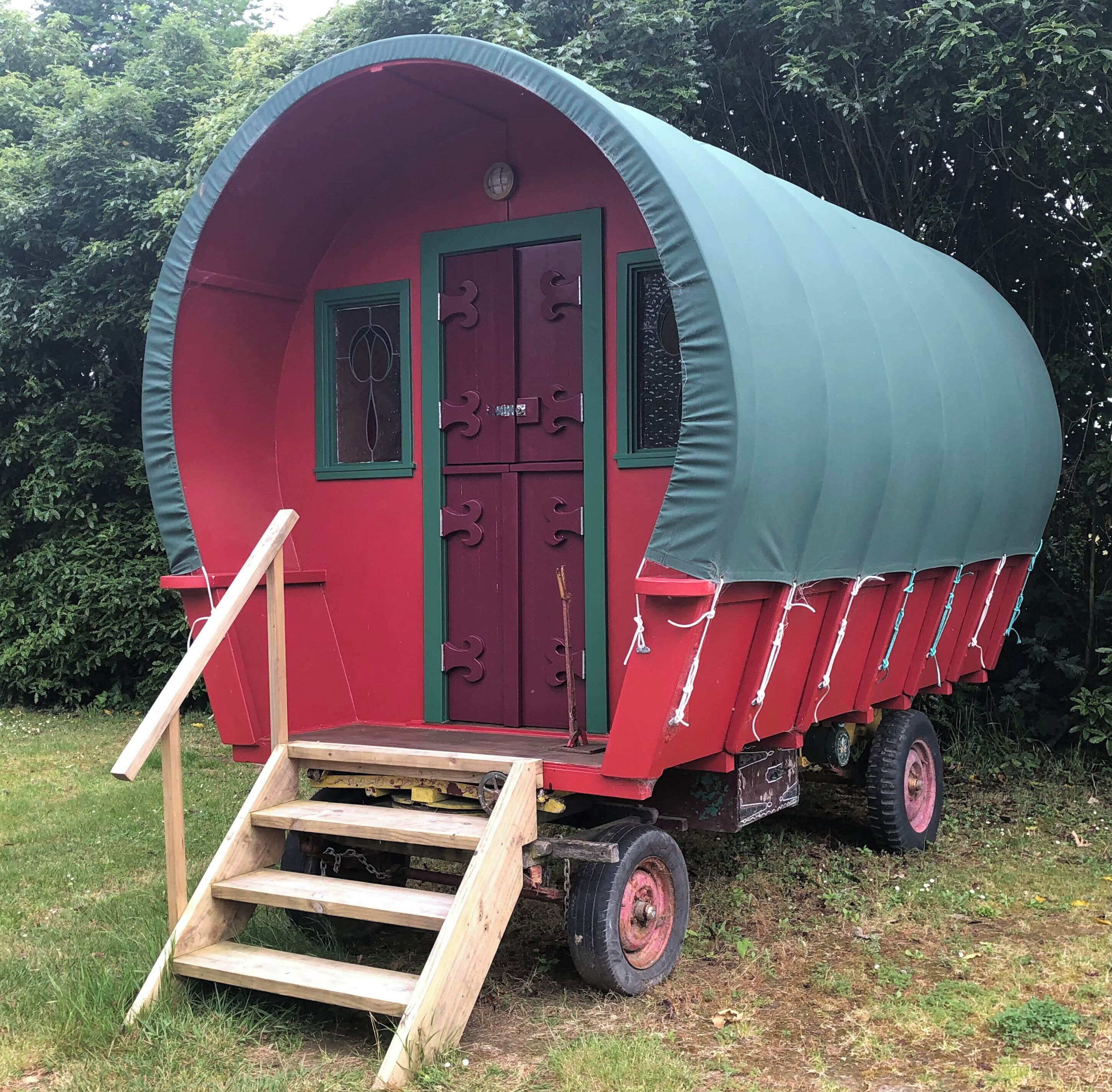A covered trailer with a red exterior and a green roof, featuring a set of wooden steps leading to a door with a window.
