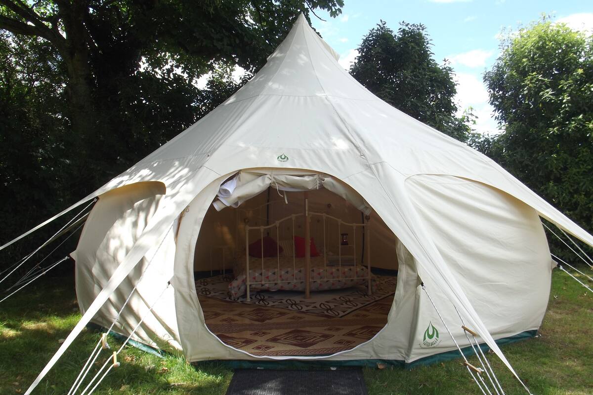 A dome-shaped tent with a bed inside, surrounded by grass and trees.