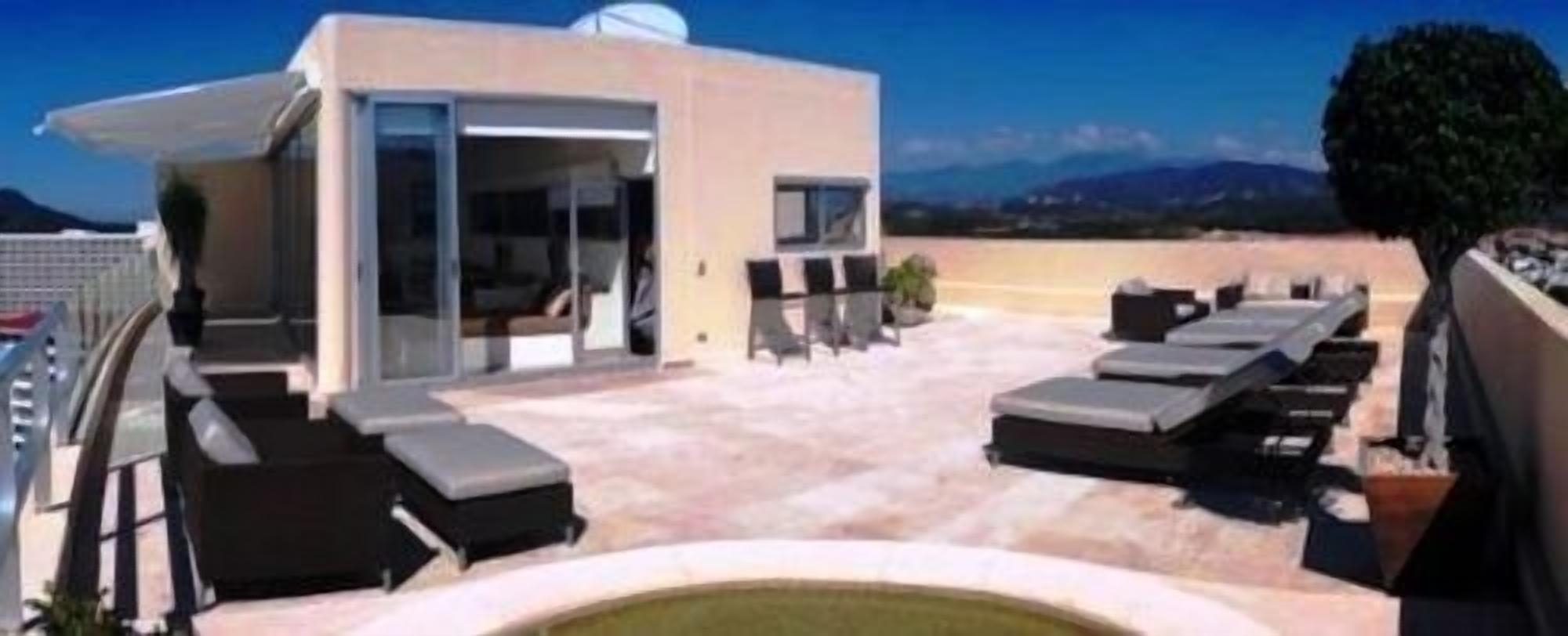 A rooftop terrace with sun loungers, a pool, and a modern building with large windows.