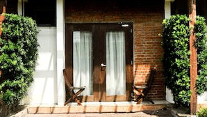 Un bâtiment à deux étages avec une façade en briques, une pergola en bois au-dessus de l’entrée, deux chaises en bois et une porte avec des rideaux.