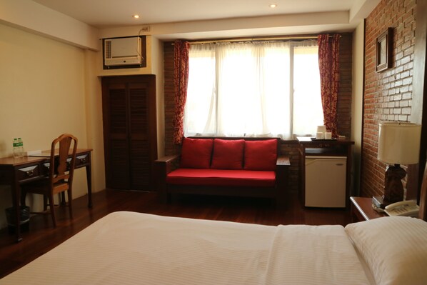 Una habitación de hotel con cama, sofá rojo, escritorio con silla, refrigerador pequeño y ventana con cortinas.