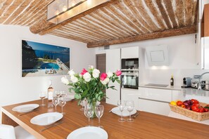 A dining area with a wooden table set for six, a vase of flowers, and a kitchen in the background.