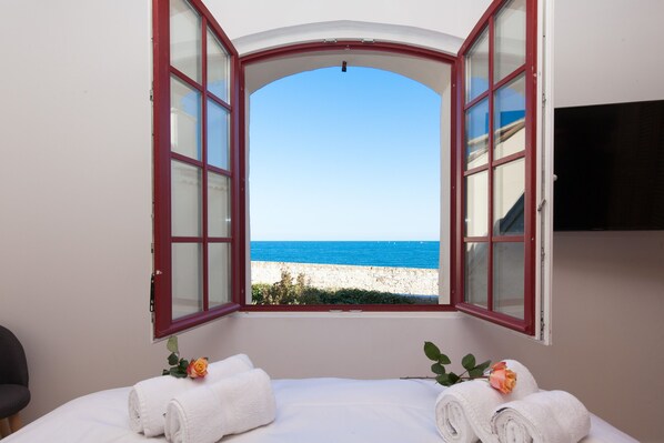 A room with a bed, two rolled towels, a rose, and a view of the sea through an open window.