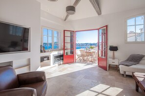 A spacious living area with a large TV, a dining table with chairs, a sofa, and a red-framed door leading to a balcony with a view of the sea.