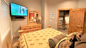 Quarto com cama, cômoda, televisão, espelho, porta e um quadro emoldurado na parede.