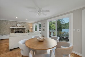 A modern living room with a round wooden table, white sofas, a stone wall, and a view of a lake. - Lake Sunapee Waterfront, Sandy Beach Cove, 75' Dock & minutes from Mt. Sunapee (Newbury)