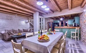 Un coin repas accueillant, avec une table dressée pour quatre personnes, entourée de chaises en bois. On aperçoit également un banc en bois et un canapé. La pièce est agrémentée de poutres apparentes au plafond et de murs en pierre.