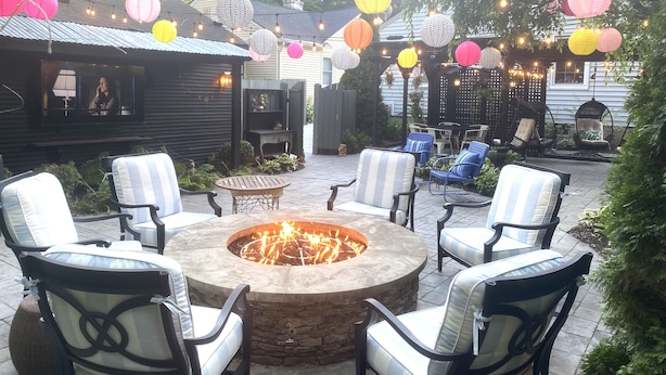 Un espace extérieur aménagé en patio, avec un foyer en pierre entouré de chaises et de tables, de guirlandes lumineuses et de lanternes suspendues.