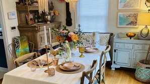Un coin repas avec une table en bois dressée pour quatre personnes, entourée de chaises. Deux lustres sont suspendus au-dessus de la table, et une fenêtre avec des stores se trouve à droite.