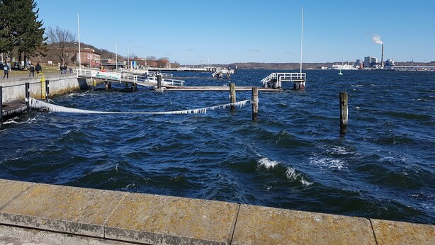 Une jetée bordant le rivage, avec des quais, un ciel dégagé et une usine industrielle visible au loin.