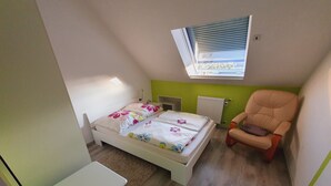 Une chambre à coucher comprenant un lit, une chaise, une petite table, un radiateur et une fenêtre avec des stores.