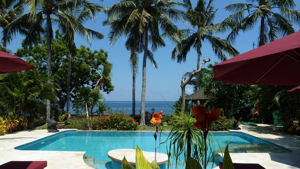 Uma área de piscina com vista para o oceano, rodeada por palmeiras e guarda-sóis vermelhos.
