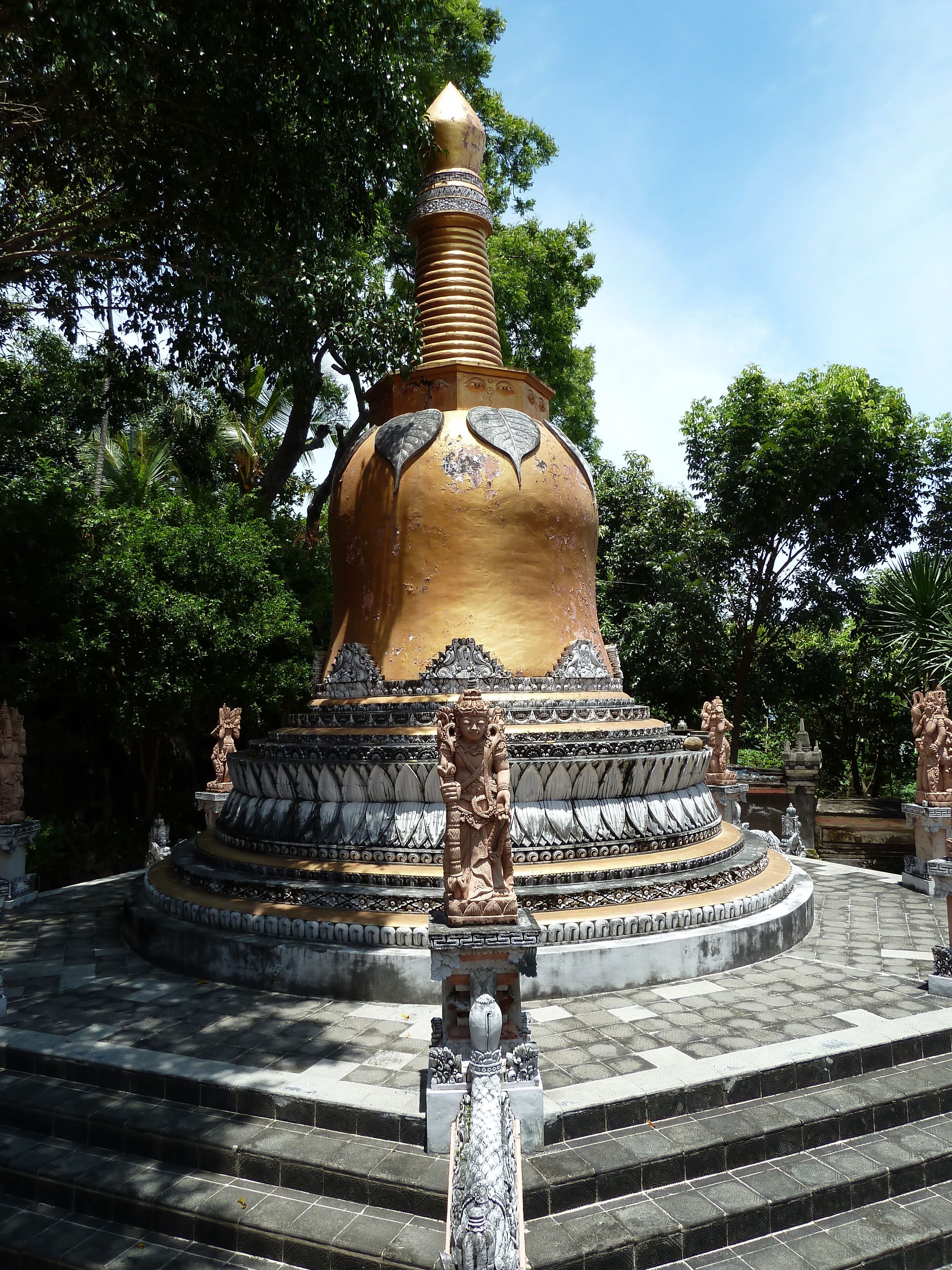 Una gran estupa dorada con intrincados relieves, rodeada de estatuas y escalones que conducen hacia ella.