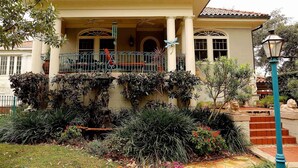 Une maison à deux étages dotée d’un balcon, de colonnes et d’un jardin où poussent différentes plantes, ainsi qu’une statue.