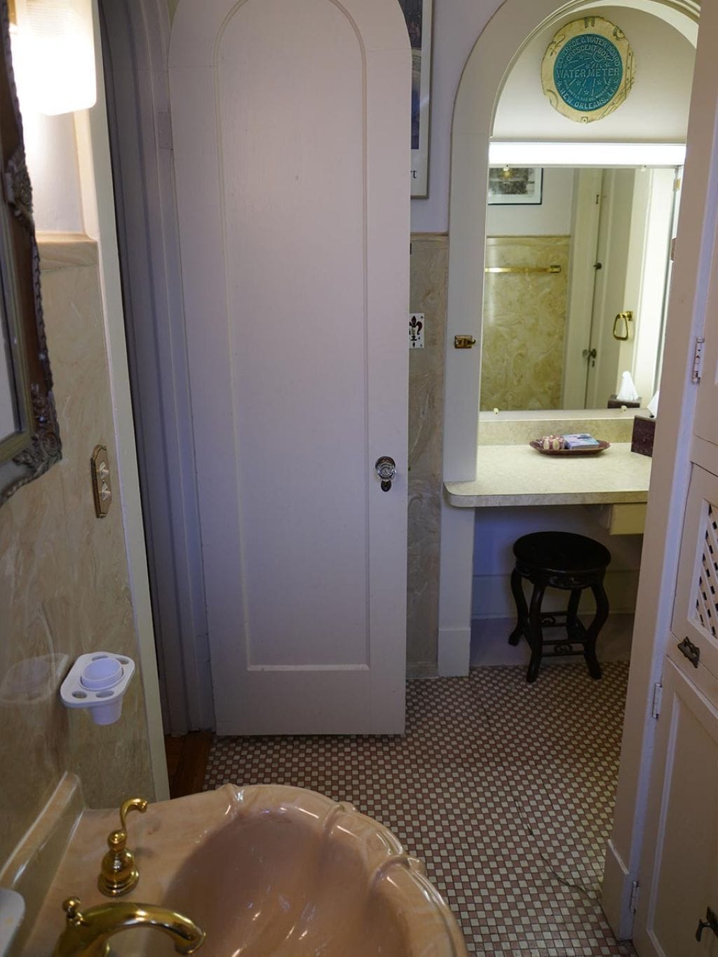 Une salle de bain avec une porte blanche, un lavabo avec un robinet doré, un petit tabouret et un miroir au-dessus duquel se trouve un compteur d’eau.