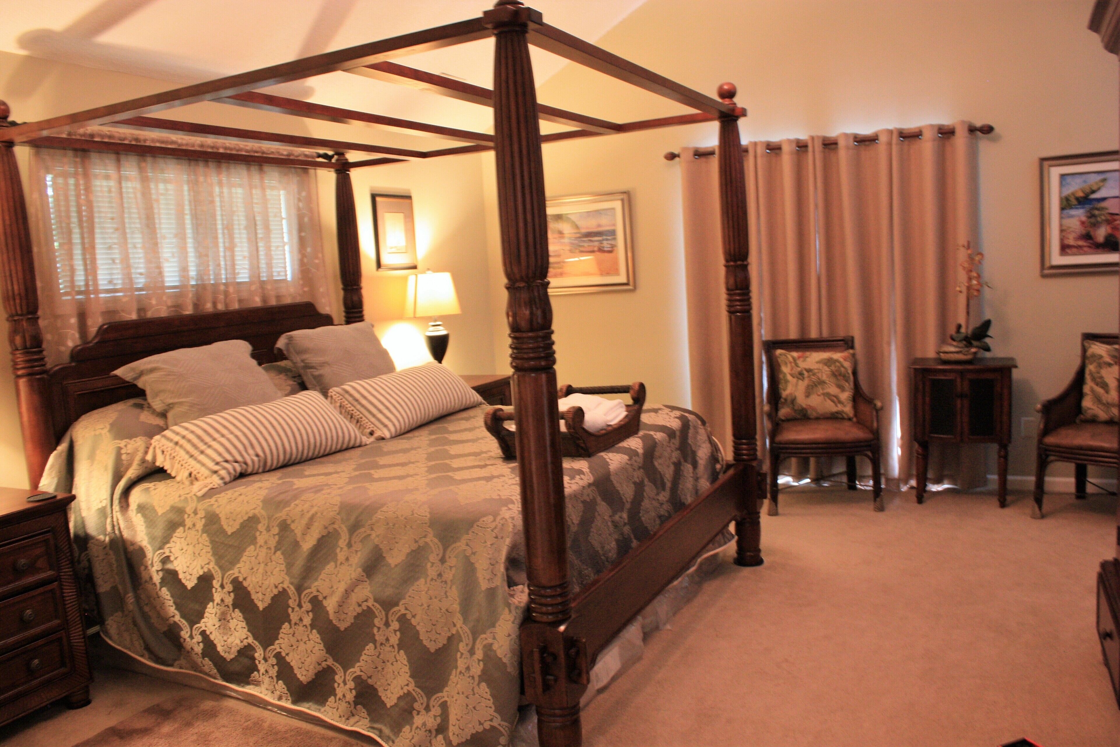 A four-poster bed with a wooden frame, a bedside table, two chairs, a small table with a plant, and a lamp.