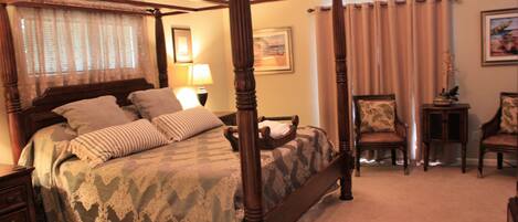 A four-poster bed with a wooden frame, a bedside table, two chairs, a small table with a plant, and a lamp.