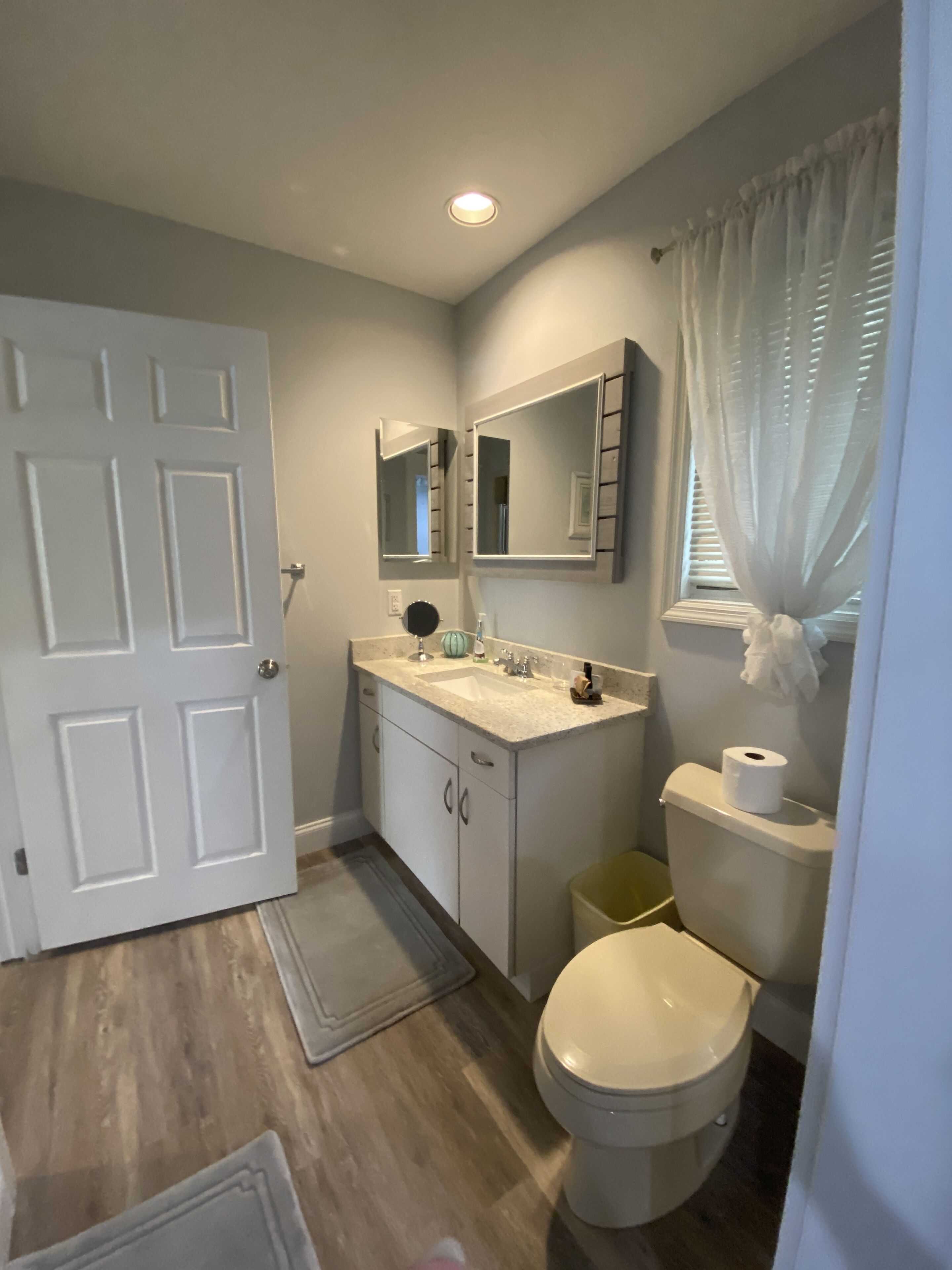 A bathroom with a white door, a sink with a mirror above it, a toilet, and a trash bin.