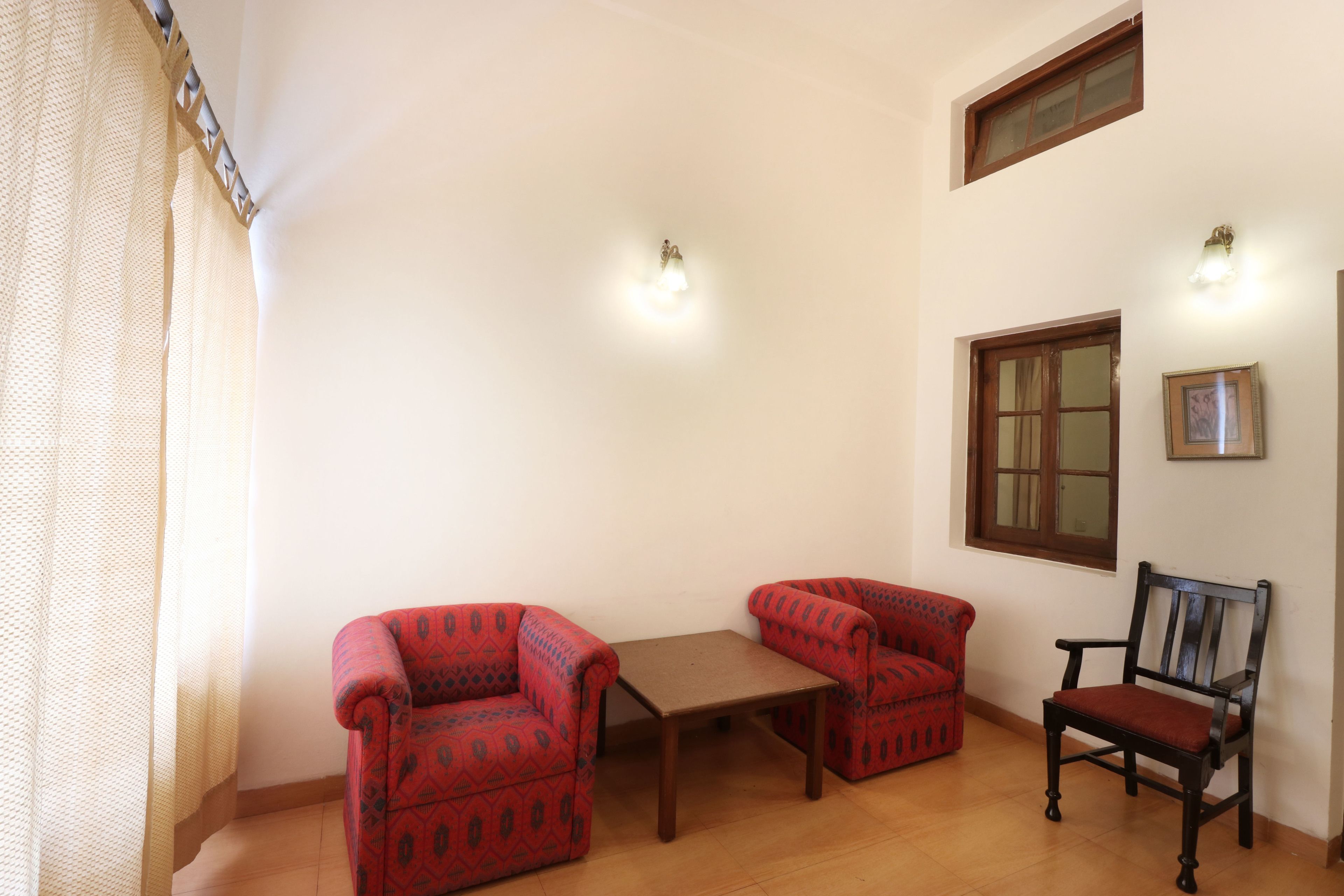 A room with two patterned armchairs, a wooden table, and a wooden chair. There are two wall-mounted lights and a window with a wooden frame.
