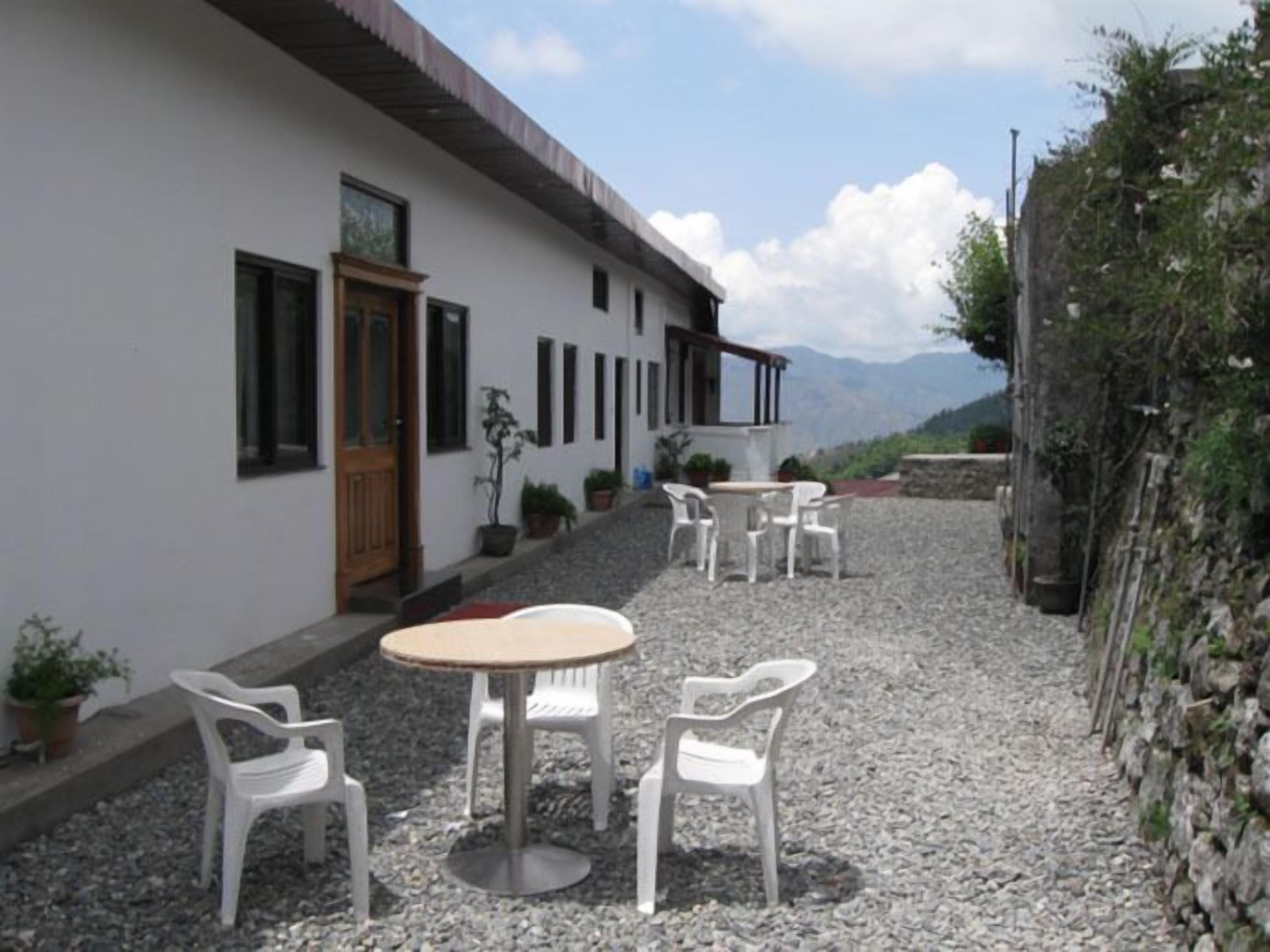 A building with a white exterior, a wooden door, and a balcony. There are three white plastic chairs and a round table in front of the building. The ground is covered with small rocks, and there are potted plants on the steps.