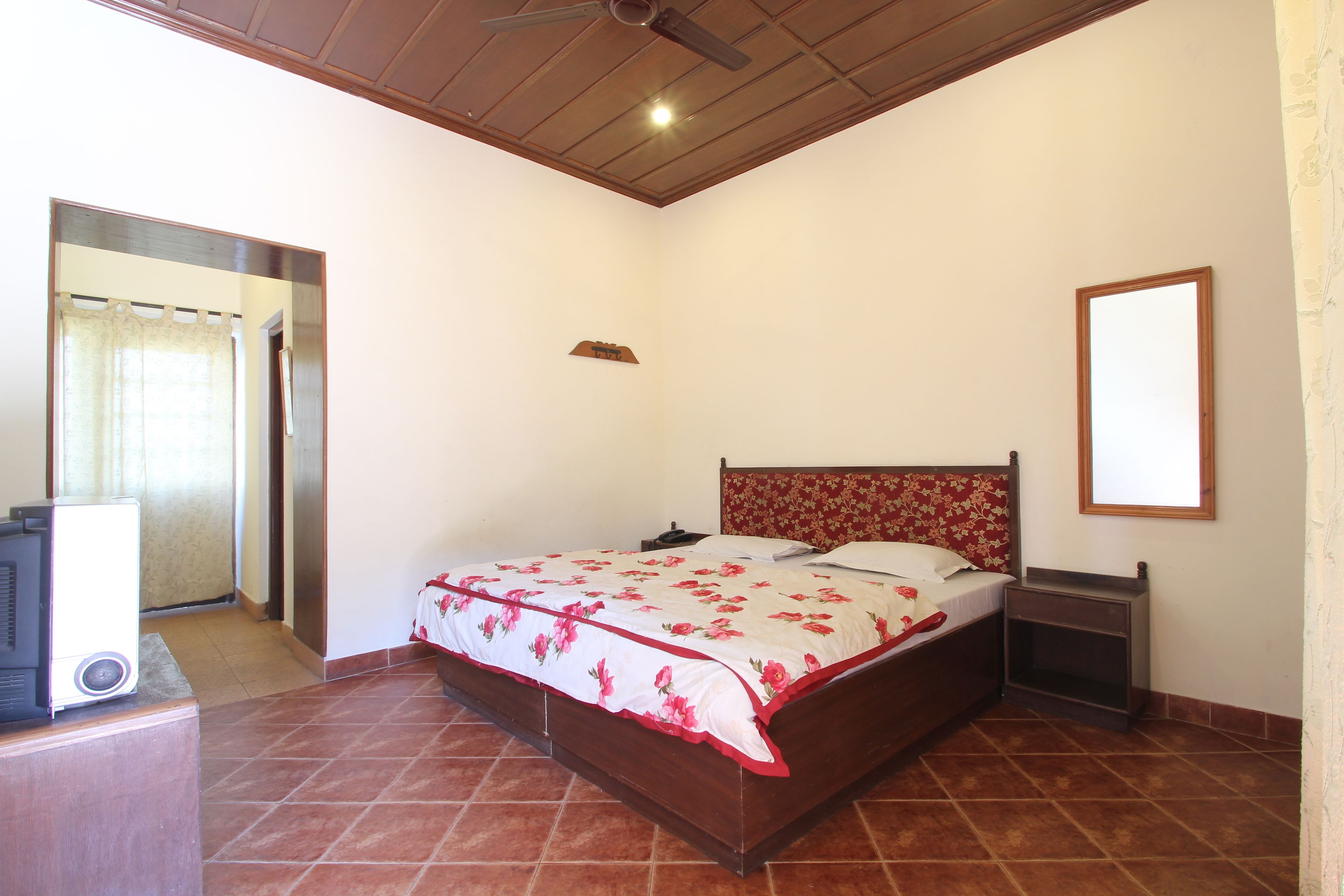 Un dormitorio con una cama de madera, una mesita de noche, un ventilador de techo y un baño a la vista a través de una puerta abierta.