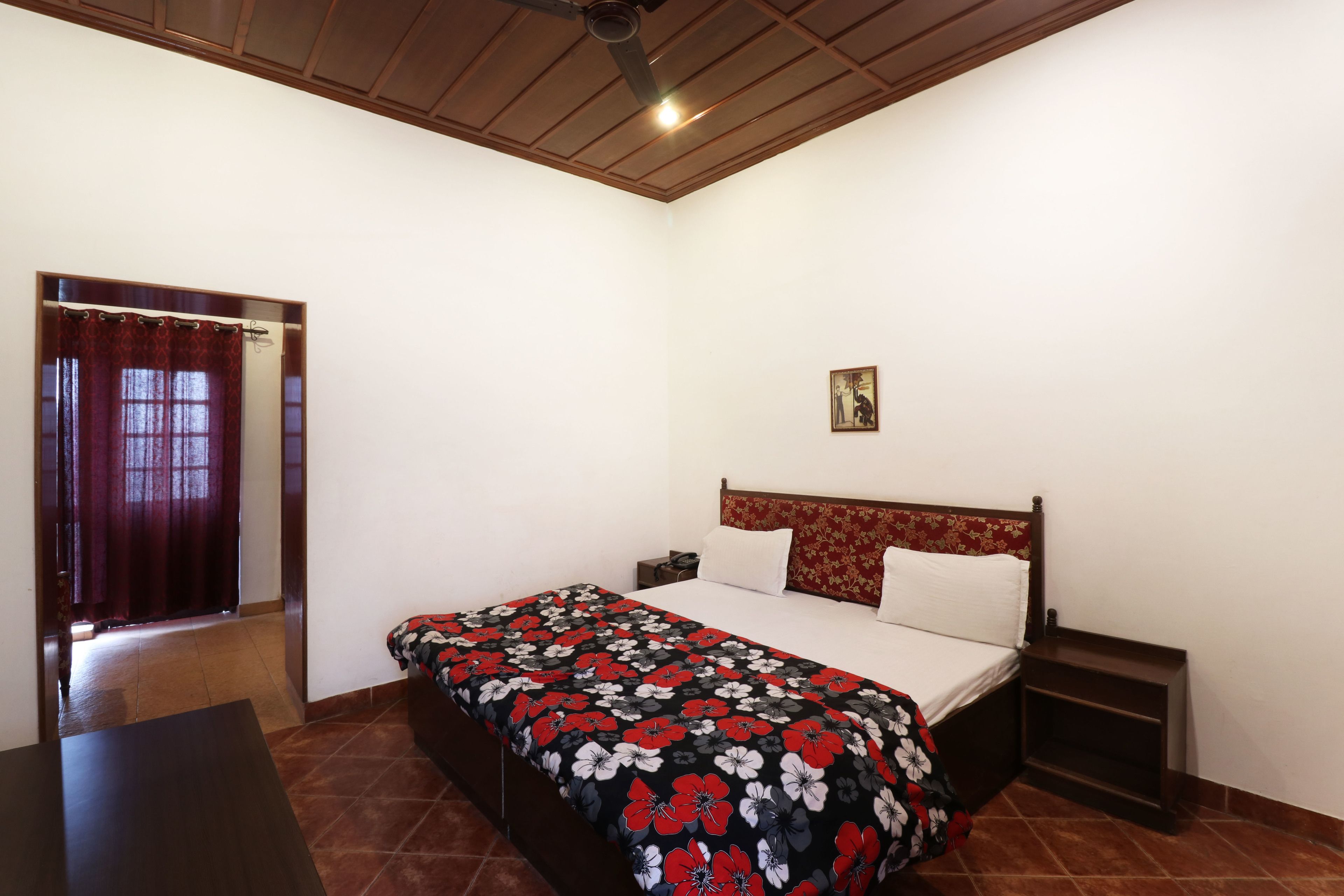 A bedroom with a bed, a nightstand, a wooden headboard, a small table, and a window with curtains.