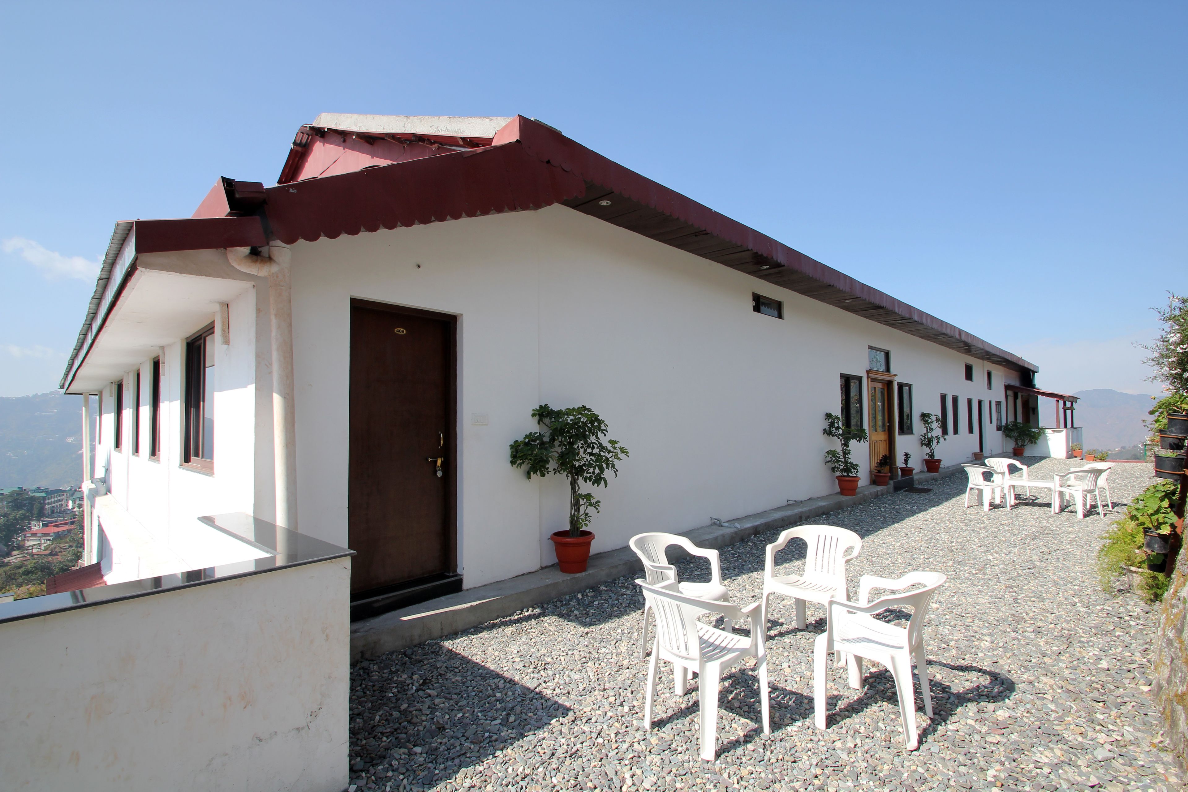 A building with a red roof and white walls, a door, and several potted plants. There are white plastic chairs and tables outside.