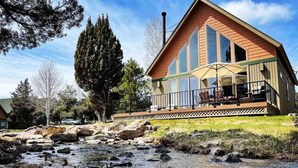 A lodge with a deck overlooking a rocky stream, large windows, and a prominent chimney. - Cozy knotty pine chalet with creekside deck, BBQ, and access to pools, hot tubs, gym, golf, hiking. (Redmond)