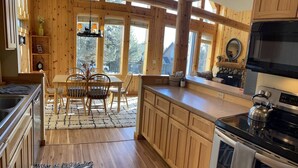 A spacious kitchen with wooden cabinets, a dining area with a table and chairs, and large windows allowing natural light. - Cozy knotty pine chalet with creekside deck, BBQ, and access to pools, hot tubs, gym, golf, hiking. (Redmond)