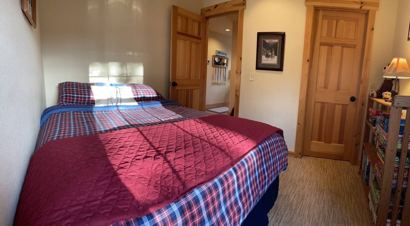 Une chambre à coucher avec un lit recouvert d’une courtepointe rouge et de draps à carreaux, des portes en bois, une bibliothèque remplie de livres et un tableau encadré accroché au mur.