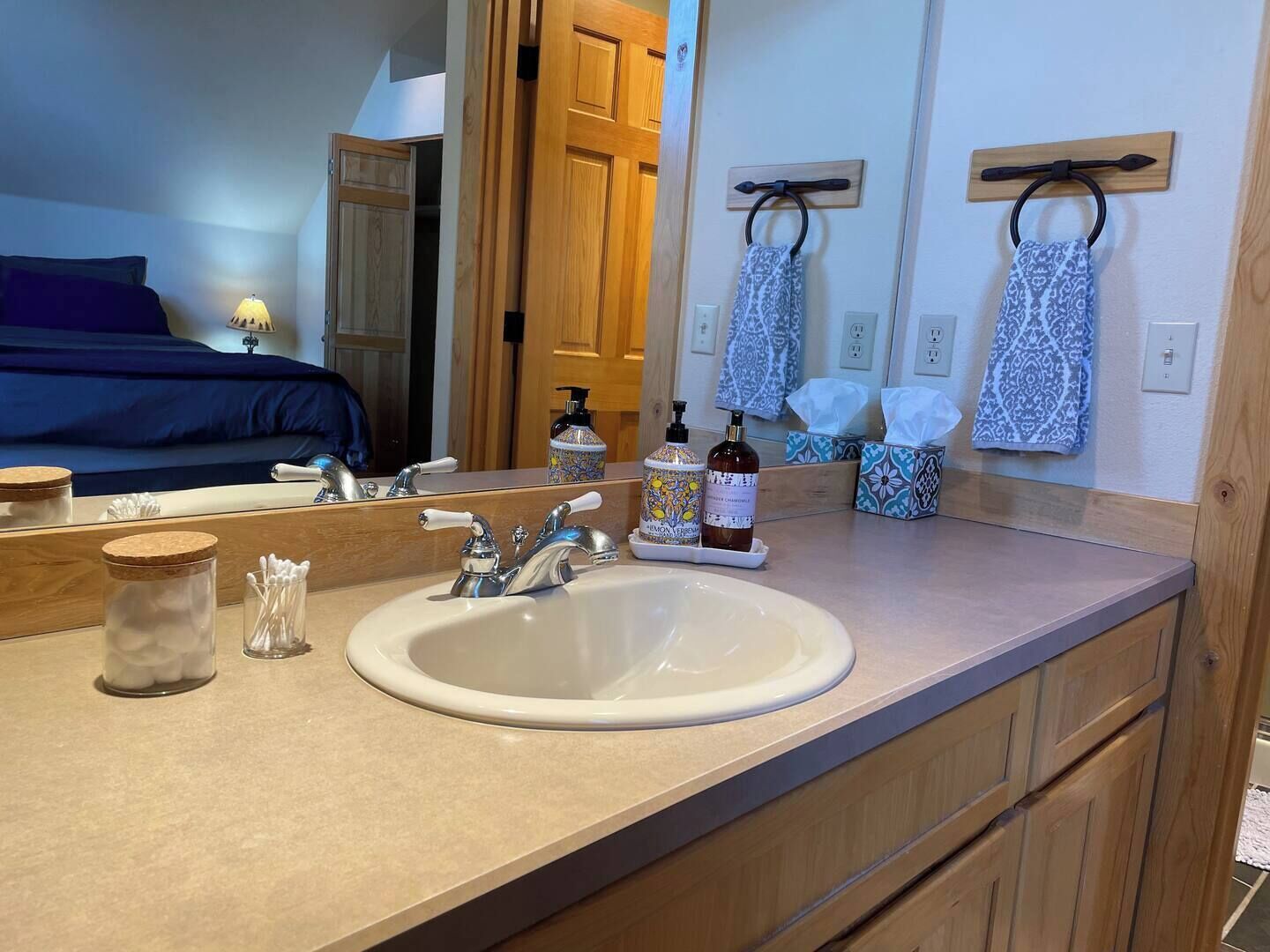 A bathroom with a large mirror, a sink, and a wooden vanity. There are two bottles of shampoo and a jar of cotton swabs on the counter.