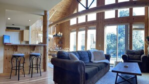 A spacious living area with a large wooden-framed window, a cozy sofa, and a coffee table. - Cozy knotty pine chalet with creekside deck, BBQ, and access to pools, hot tubs, gym, golf, hiking. (Redmond)