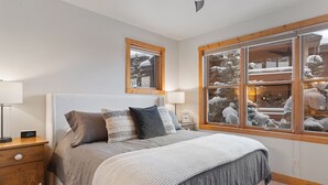 Ein Schlafzimmer mit einem großen Bett, einem Holzkopfteil, einem Nachttisch mit Lampe und einem Fenster mit Blick auf schneebedeckte Bäume.