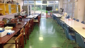 Une salle de classe avec un sol vert, des chaises et des tables en bois et bleues, un téléviseur et une façade vitrée.