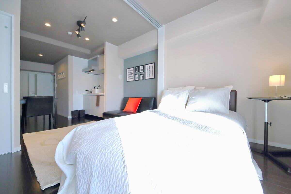 Une chambre d’hôtel moderne avec un grand lit, un bureau et une chaise. On y trouve une petite table avec une lampe et une étagère fixée au mur.