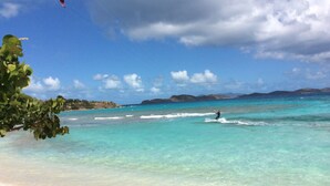 En strand med krystalklart turkis vand, en person der sejler på en windsurfingbræt, og fjerne bjerge i baggrunden.