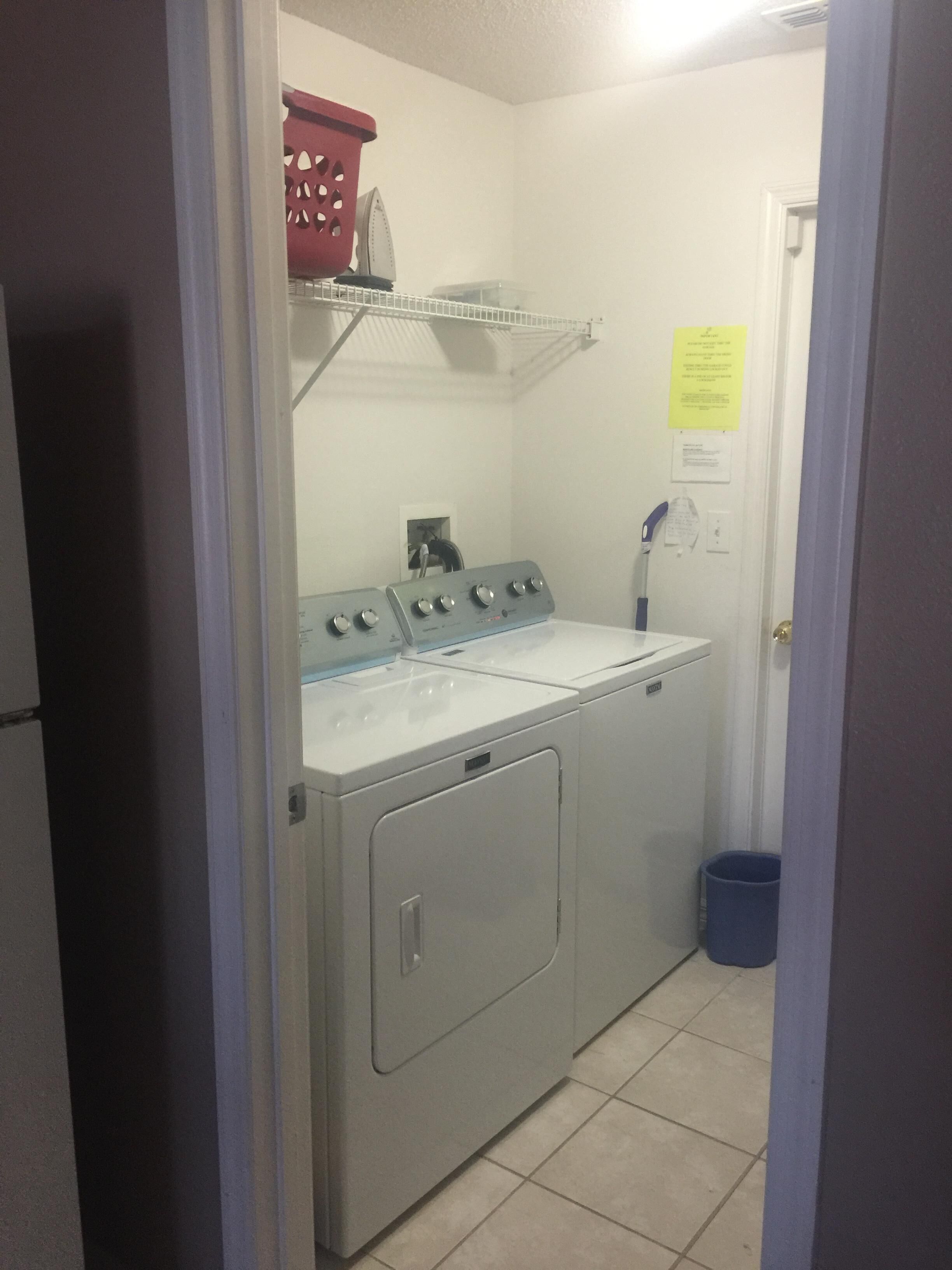 A laundry room with a front-loading washer and dryer, a red basket on a shelf, and a blue trash can.
