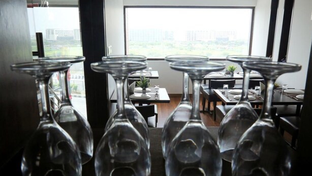 A dining area with tables set for service, a view of the city through large windows, and hanging wine glasses. - Hotel The R Blues (New Delhi)