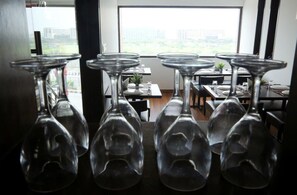 A dining area with tables set for service, a view of the city through large windows, and hanging wine glasses. - Hotel The R Blues (New Delhi)