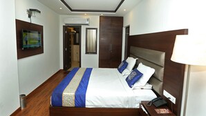 A hotel room with a bed, a desk, a telephone, a lamp, a ceiling fan, and a television mounted on the wall. - Hotel The R Blues (New Delhi)