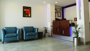 A reception area with two blue chairs, a wooden reception desk, and a potted plant. - Hotel The R Blues (New Delhi)