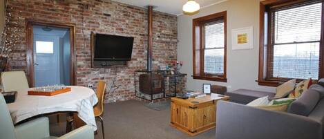A living room with a brick wall, a television mounted above a fireplace, a sofa, and a dining table with chairs.