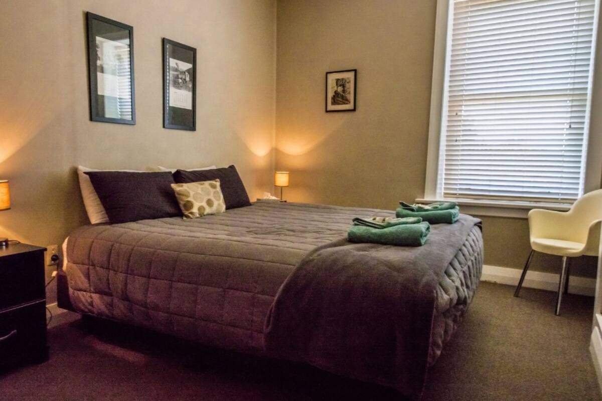 A bedroom with a bed, bedside lamps, a chair, and a window with blinds.