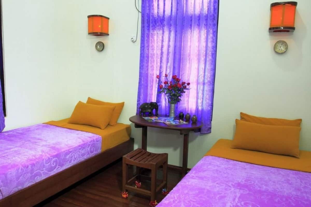 A room with two beds, each with an orange pillow and a purple bedspread. There is a small wooden stool and a table with a vase of flowers and decorative items.