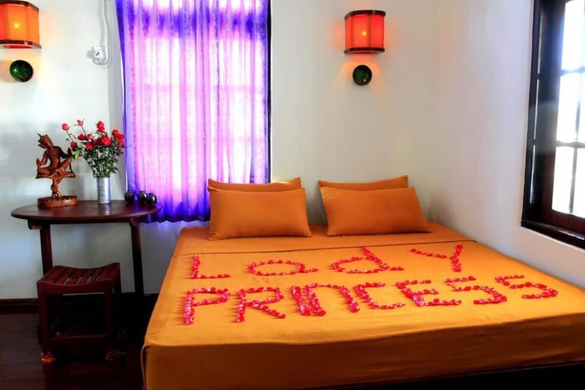 A hotel room with a bed covered in orange bedding, a decorative wooden chair, a small table with a vase of flowers, and a purple curtain.