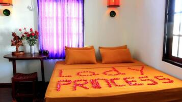 Habitación de hotel con una cama cubierta con ropa de cama naranja, una silla de madera decorativa, una mesita con un jarrón de flores y una cortina morada.