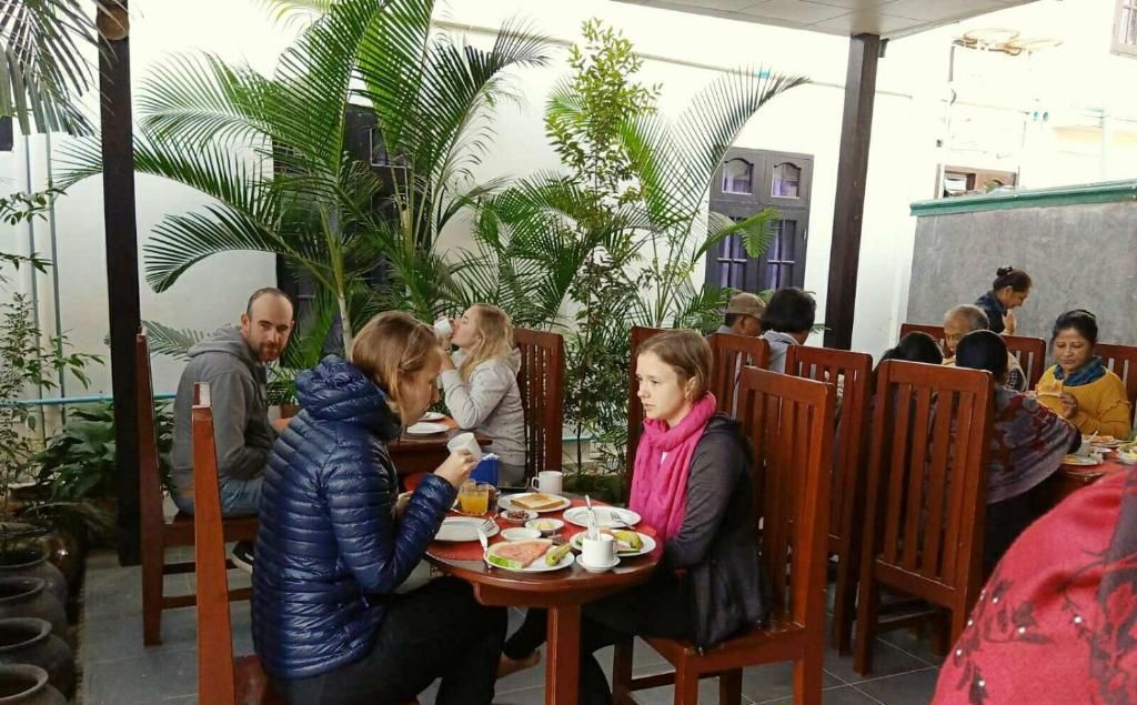 Un grupo de personas sentadas alrededor de una mesa redonda en un café al aire libre, con sillas de madera y plantas en macetas.