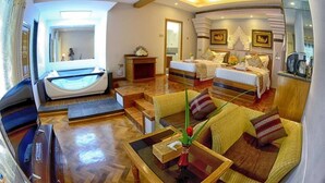 Amplia habitación de hotel con una cama grande, un área de descanso con sofá y sillones, una mesa de comedor y una pequeña cocina.