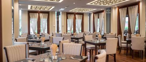 A spacious dining area with multiple tables and chairs, featuring a modern ceiling design with recessed lighting and large windows with curtains.